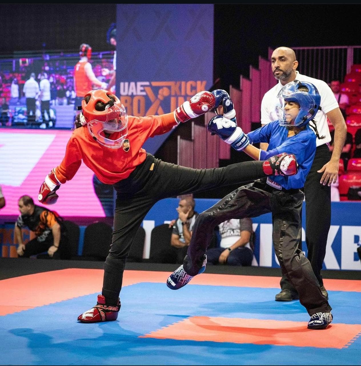 Young kickboxer in red landing a high kick on an opponent in blue in front of a UAE Kickboxing backdrop