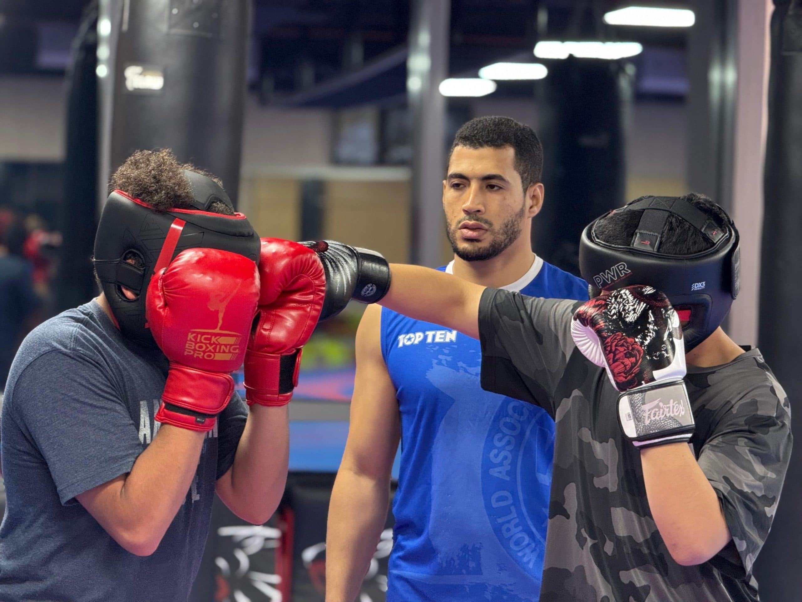 Two kickboxing students sparring in full headgear while their coach supervises