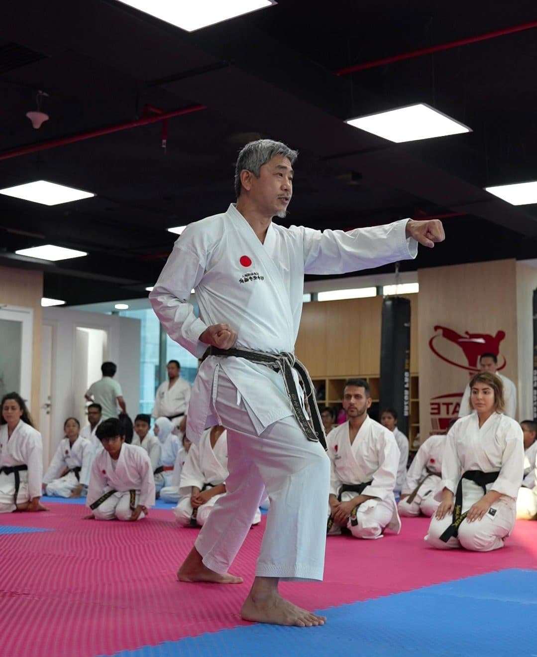 Instructor demonstrating a straight punch while seated students watch from the mats behind him