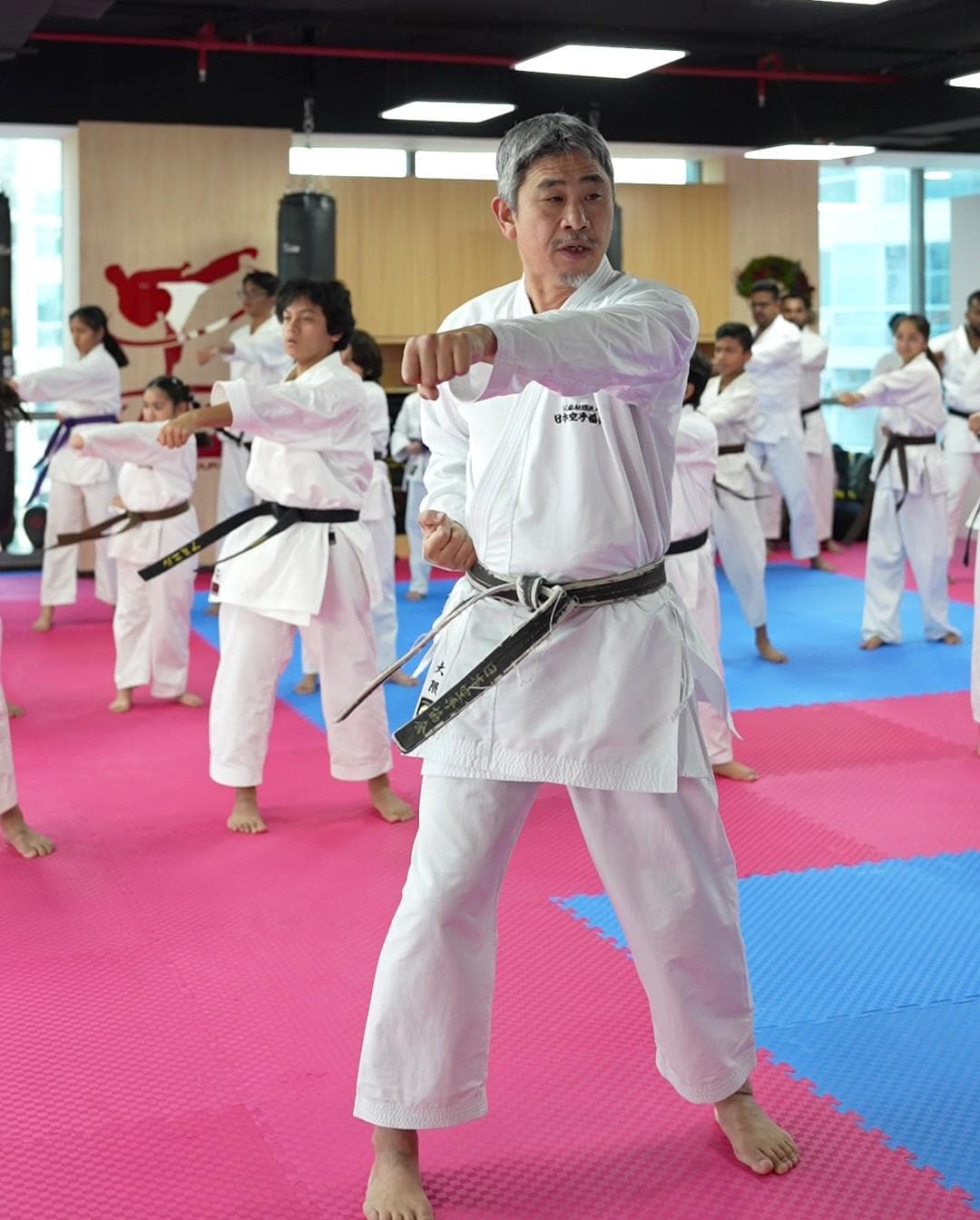 Senior instructor leading a punching drill with a class of adult karatekas spread across pink and blue mats