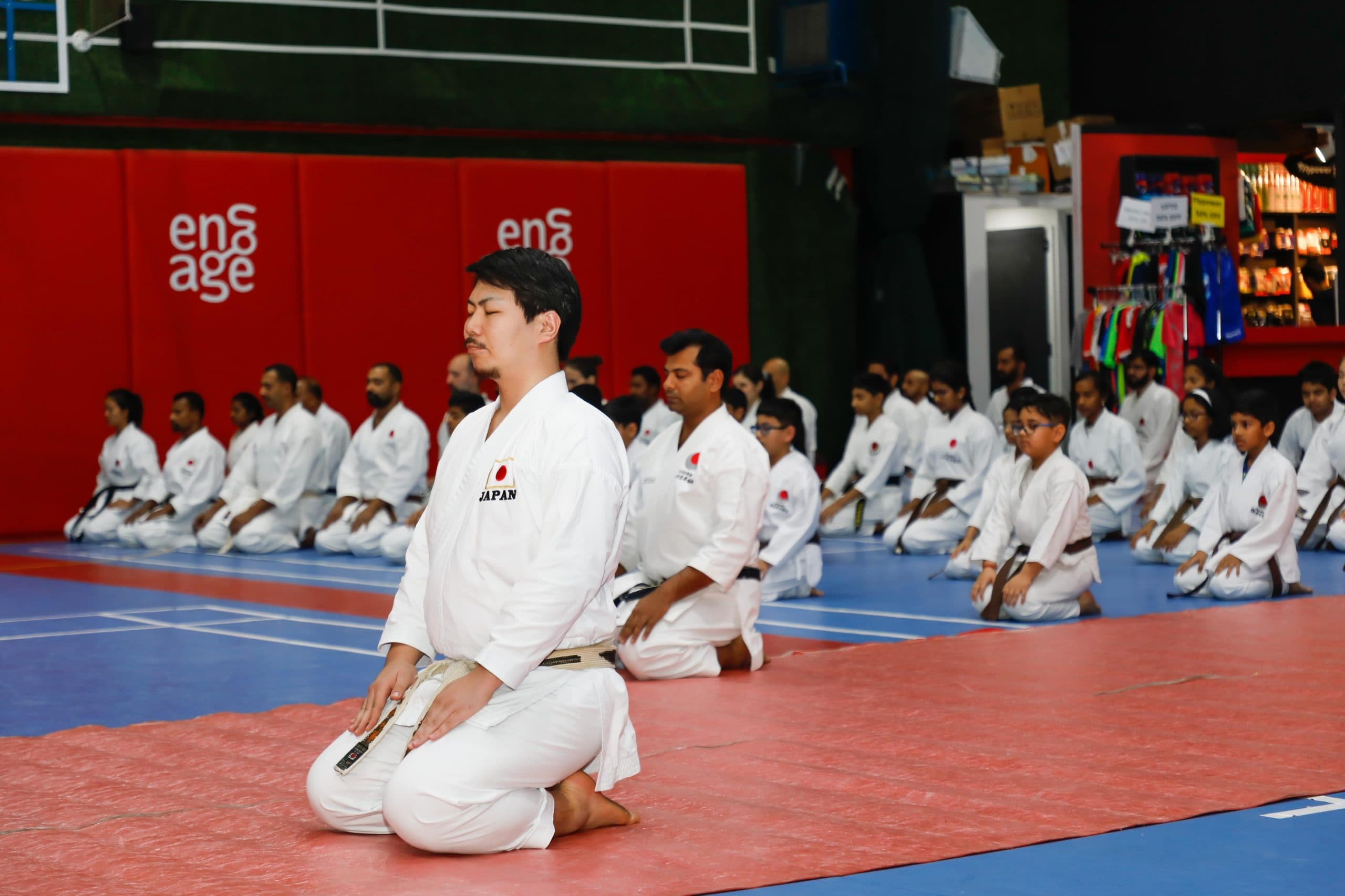 Adult karatekas kneeling in seiza in neat rows on red and blue mats in a sports hall