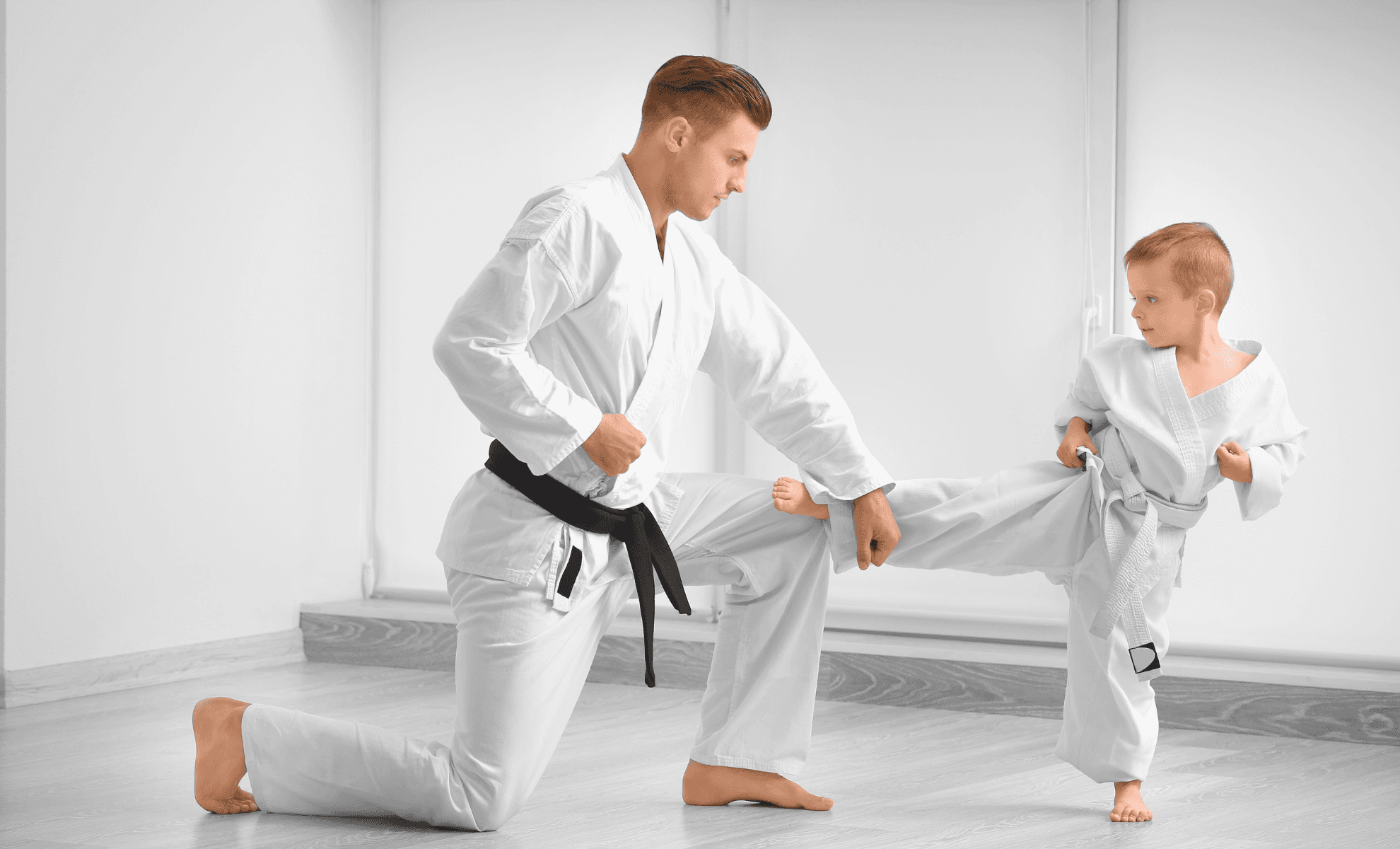 Adult black belt kneeling to coach a young student through a side kick in a white studio