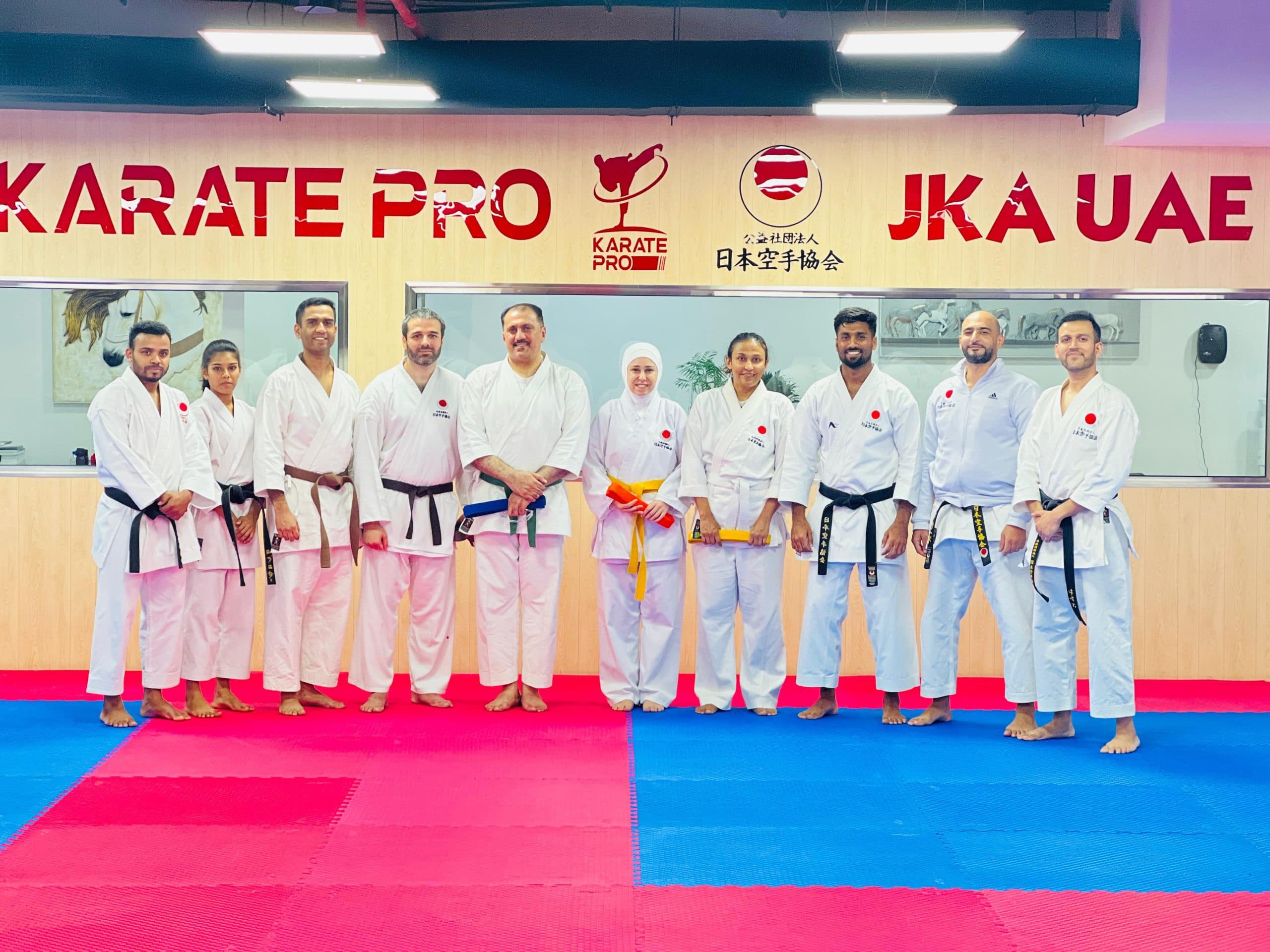 Adult karate class of mixed belt grades posing in a line beneath the Karate Pro and JKA UAE signage