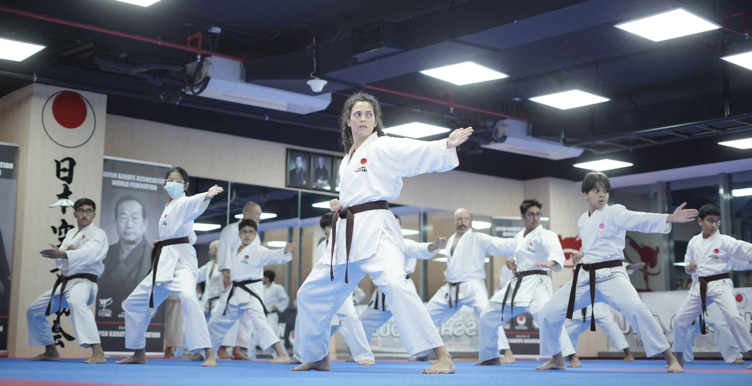 Woman in white gi extending an open-hand strike while classmates train behind her on the blue mats