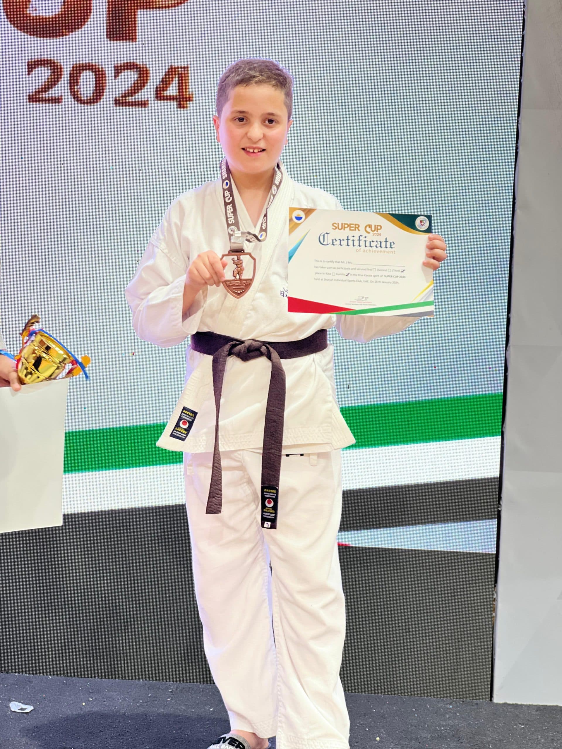 Boy in brown belt holding up his Super Cup 2024 certificate and medal on stage