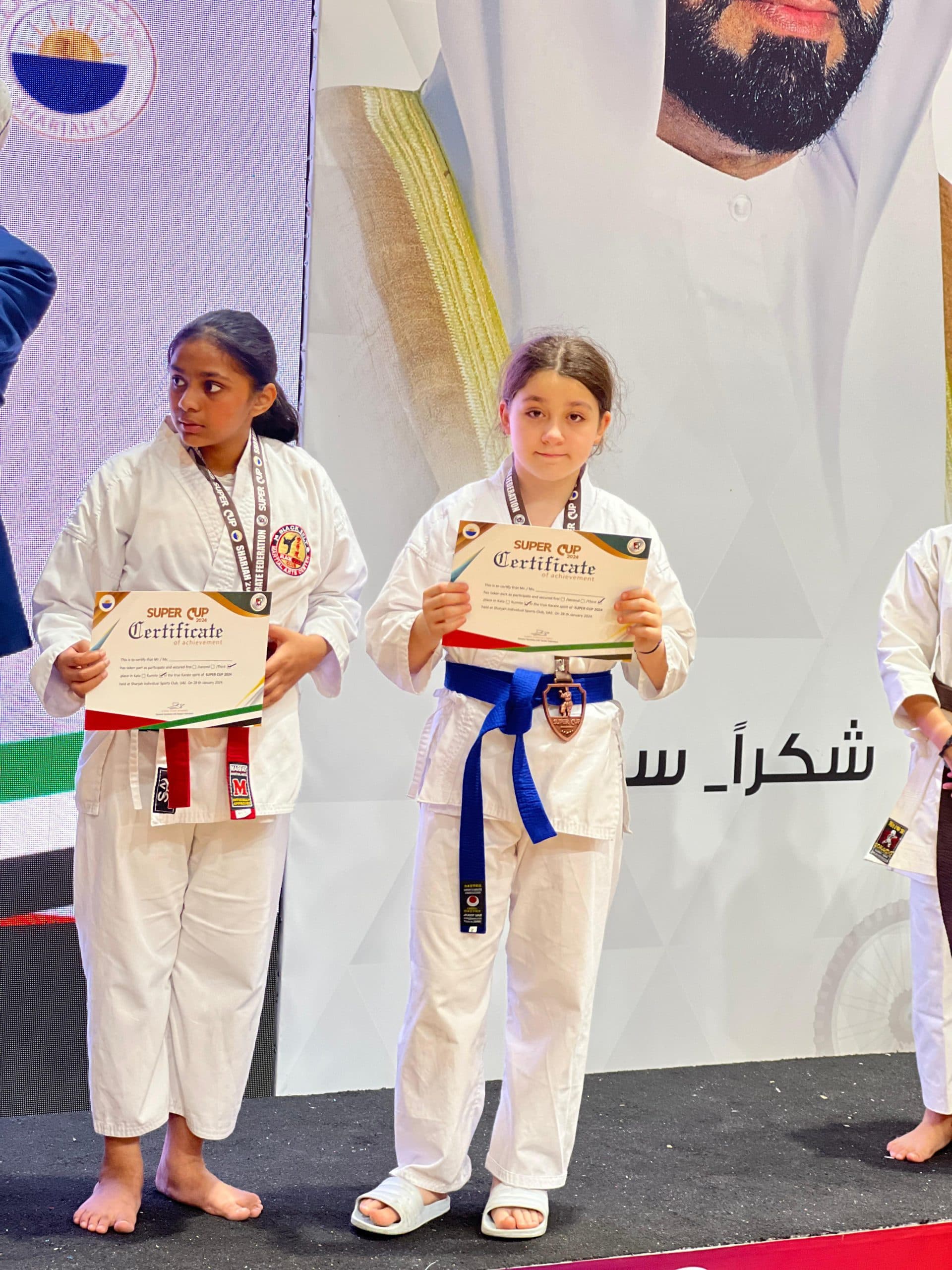 Two girls in white gi holding certificates and medals beside a wall of championship trophies