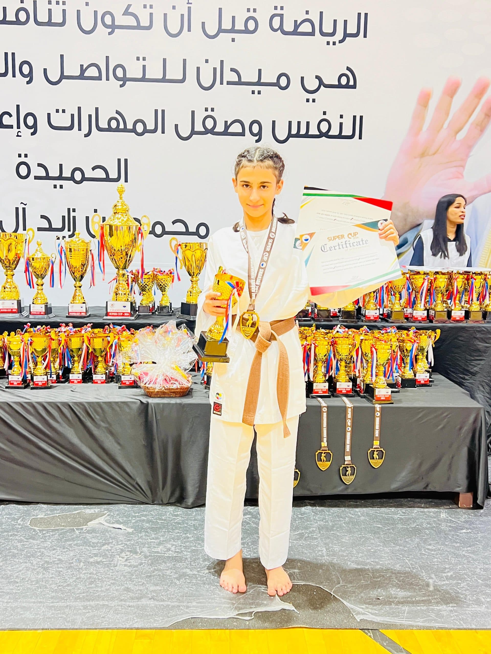 Young karateka standing with a trophy and certificate in front of tables full of championship cups