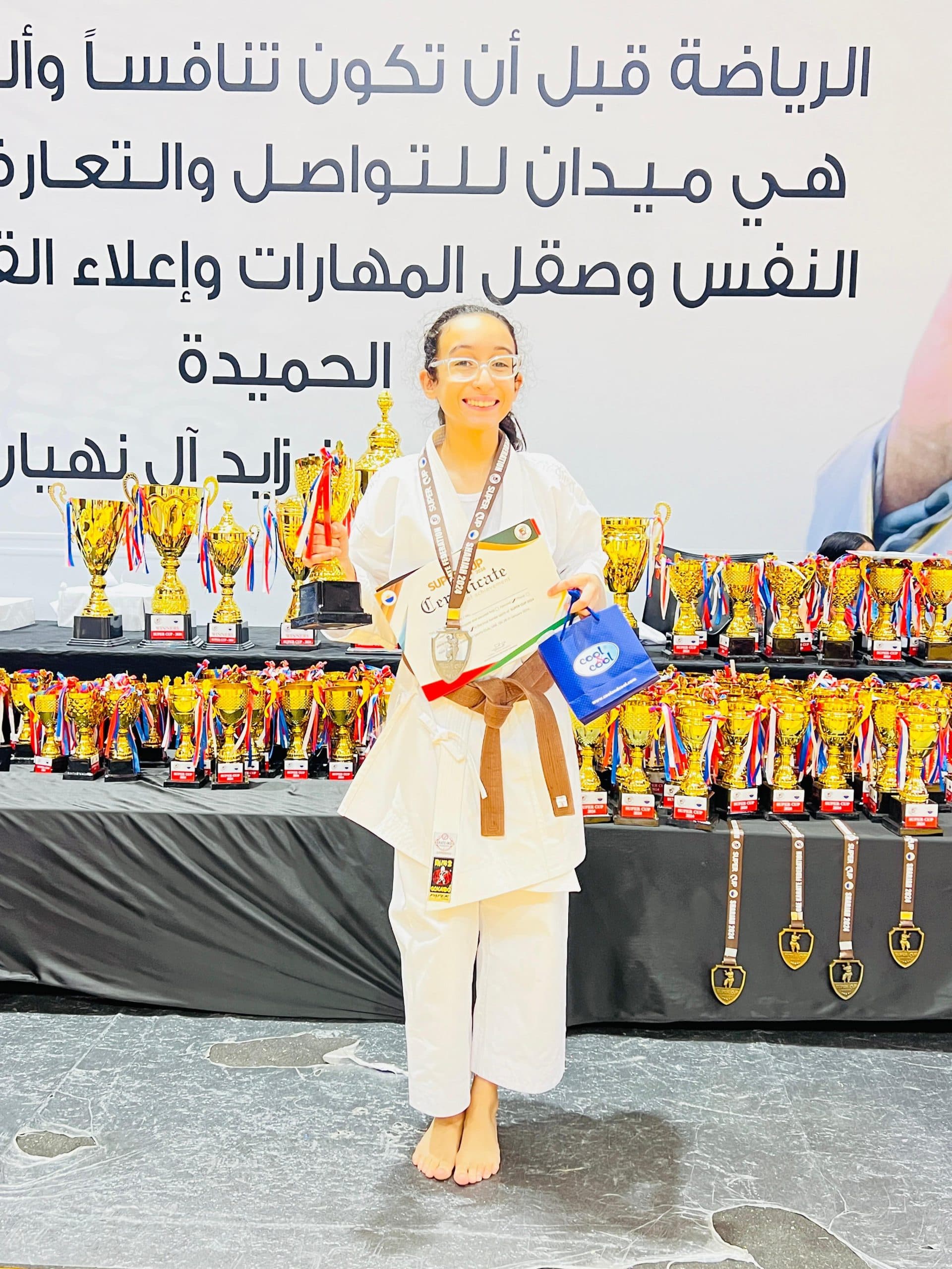 Girl in white gi with a medal holding her certificate in front of a display of trophies