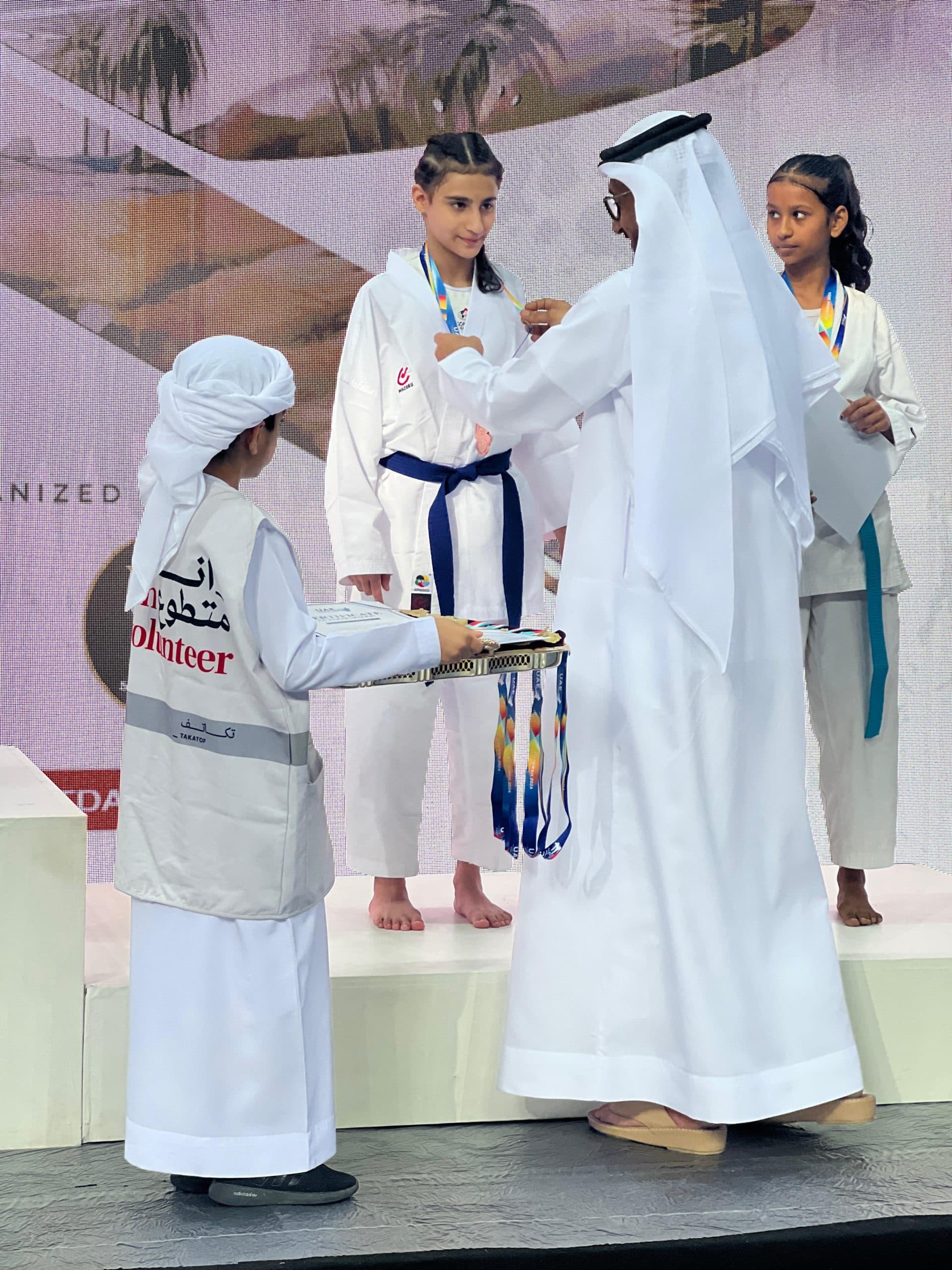 Official in traditional Emirati dress presenting a medal to a young girl in white gi on stage