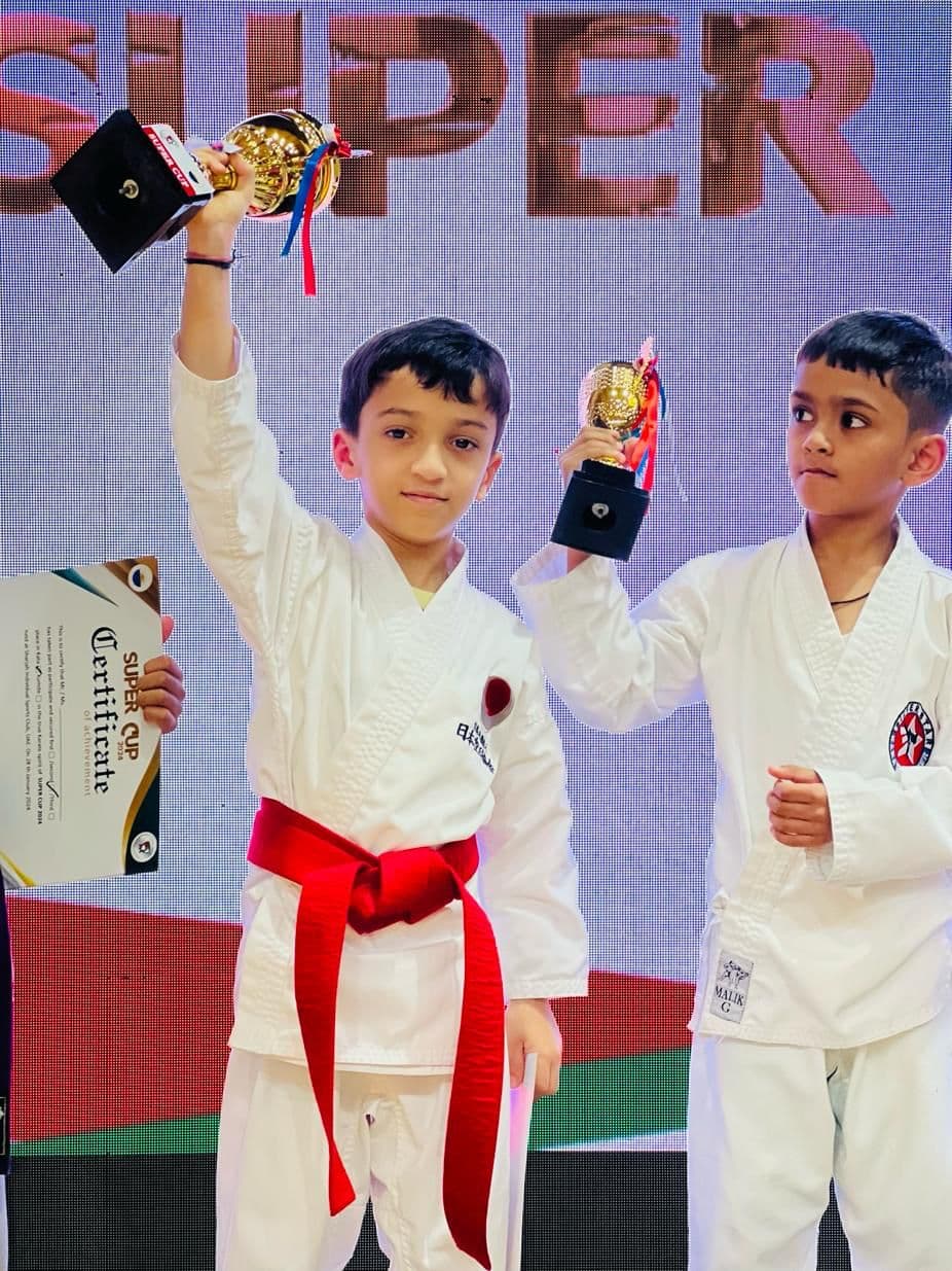 Two young boys in white gi raising trophies in celebration on stage at a Super Cup event