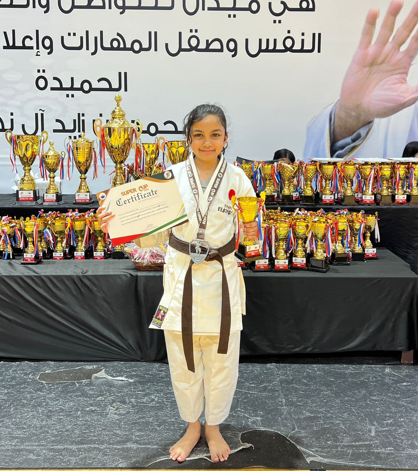 Girl in white gi with a medal and certificate standing beside a table of championship trophies