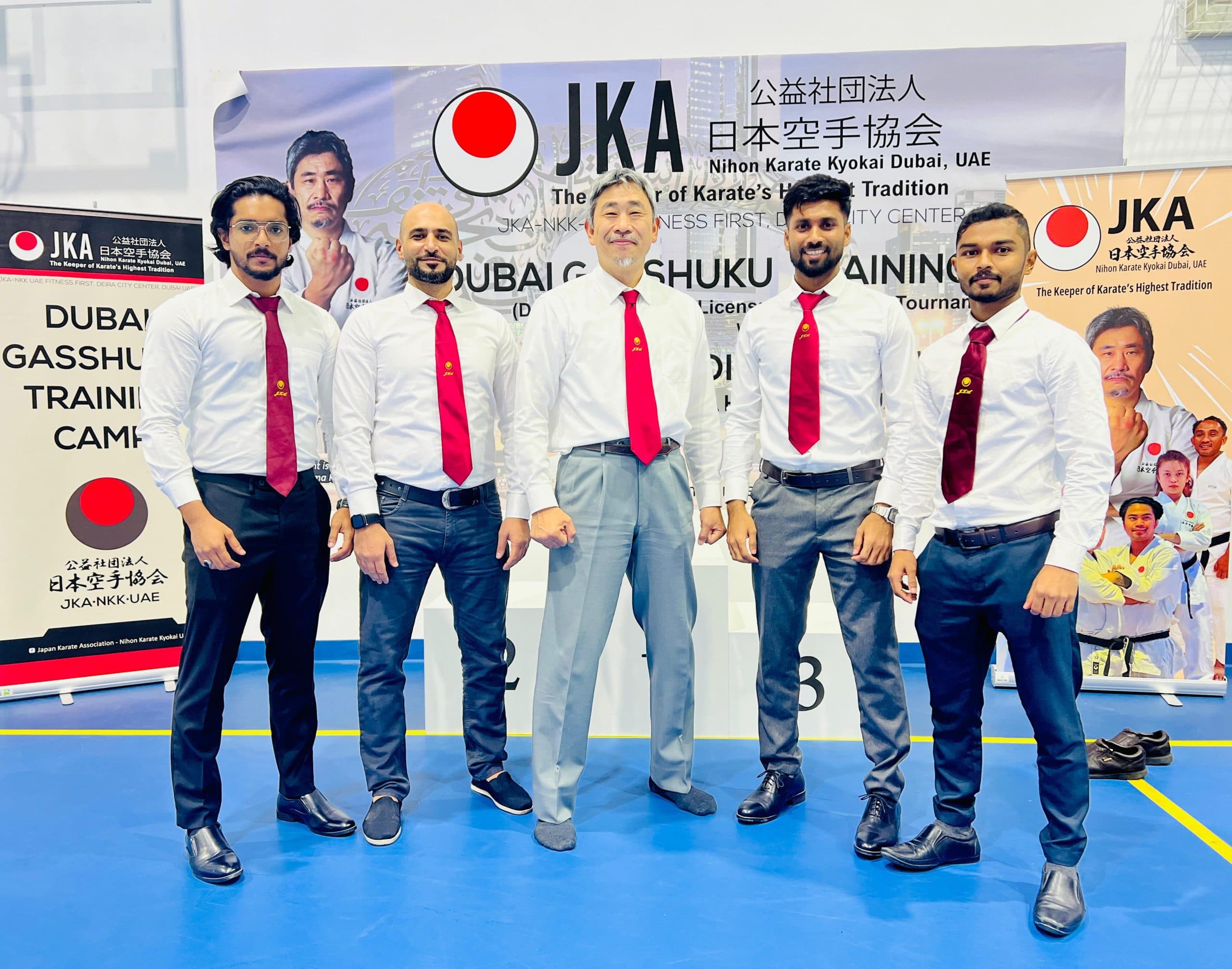 Instructors in white shirts and red ties posing in front of a JKA Dubai Gasshuku training camp banner