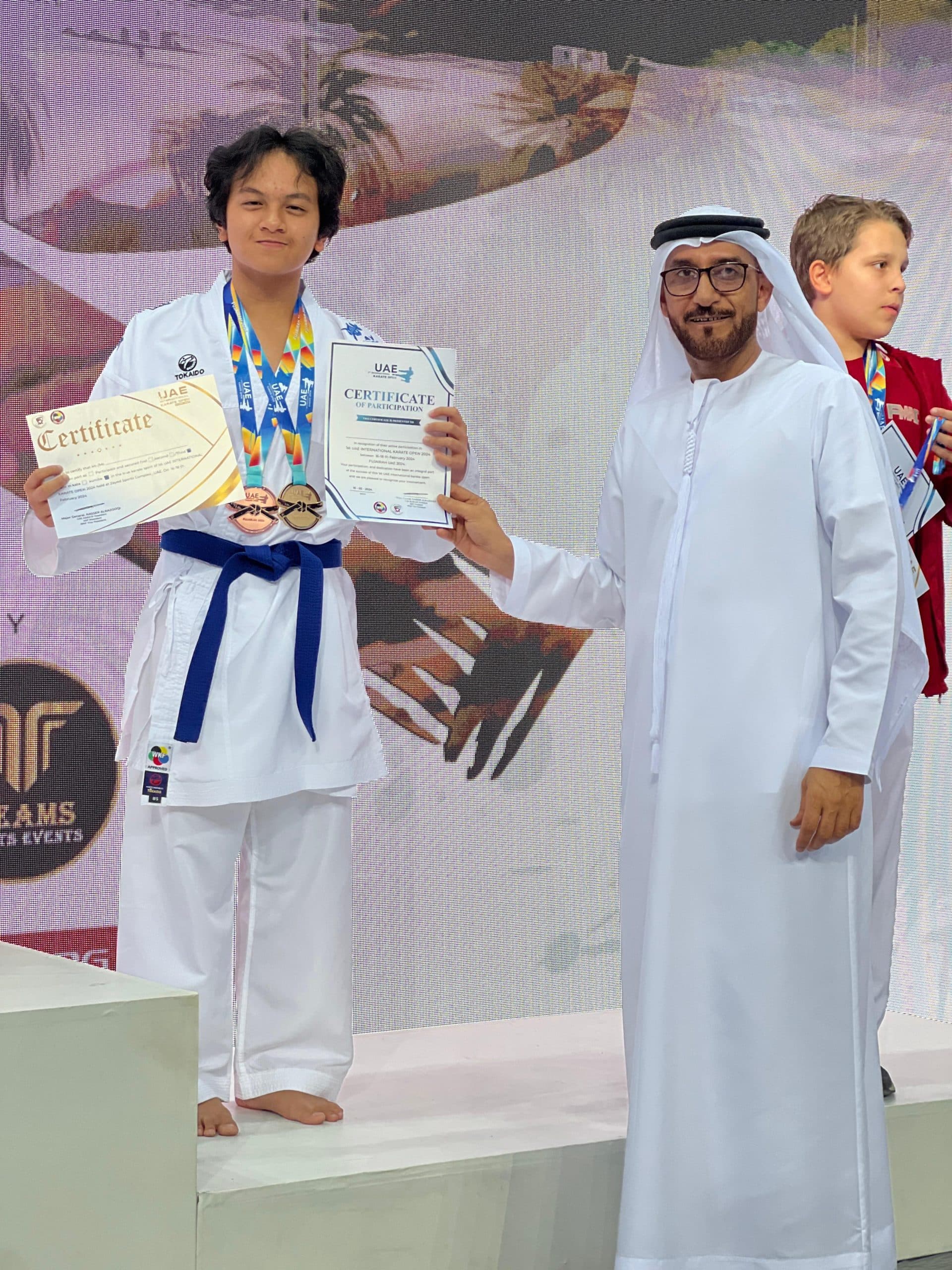 Young karateka in blue belt holding certificates and medals beside an official on the podium in Fujairah