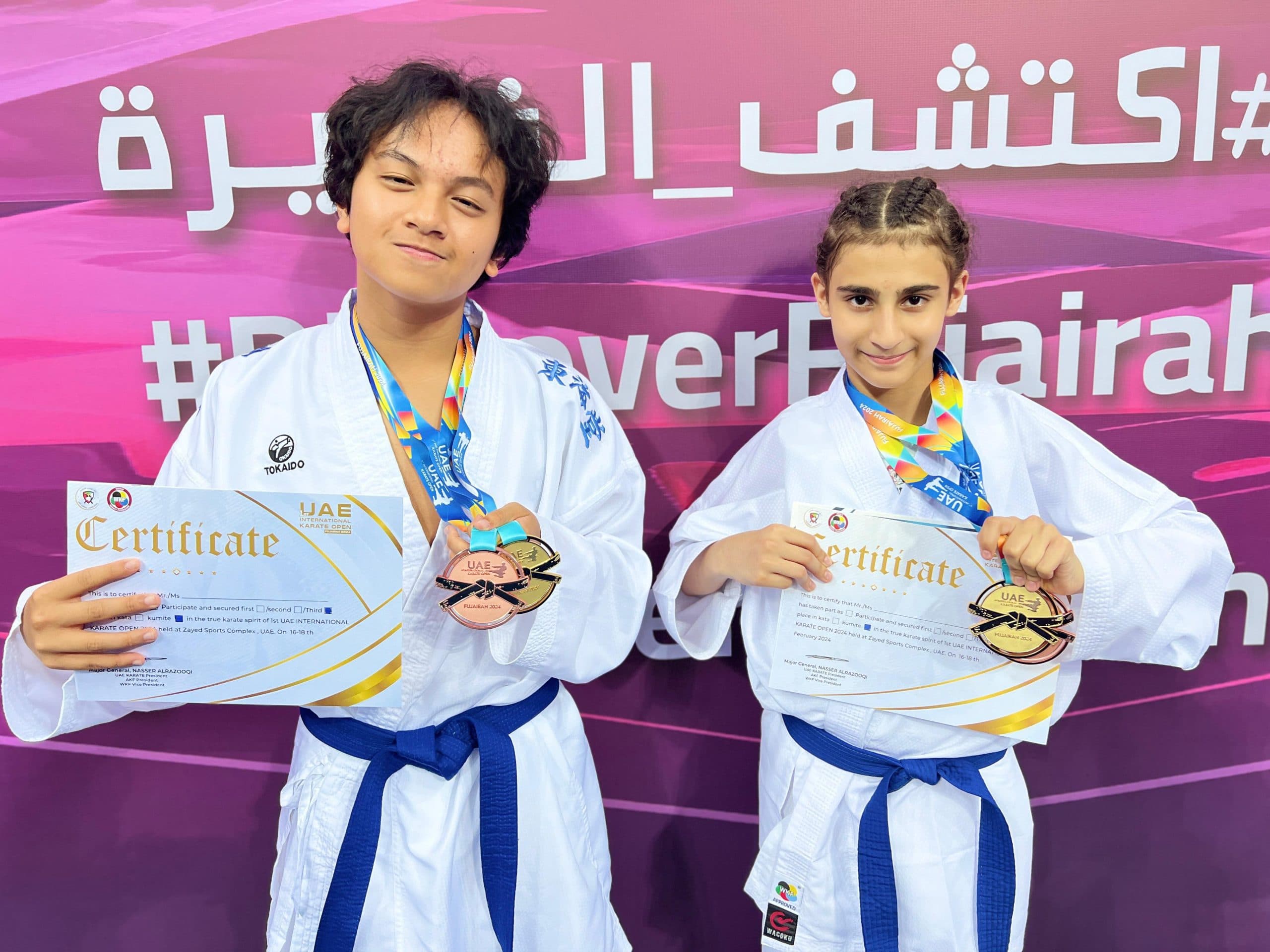 Two young competitors showing their gold medals and certificates in front of a Fujairah championship backdrop