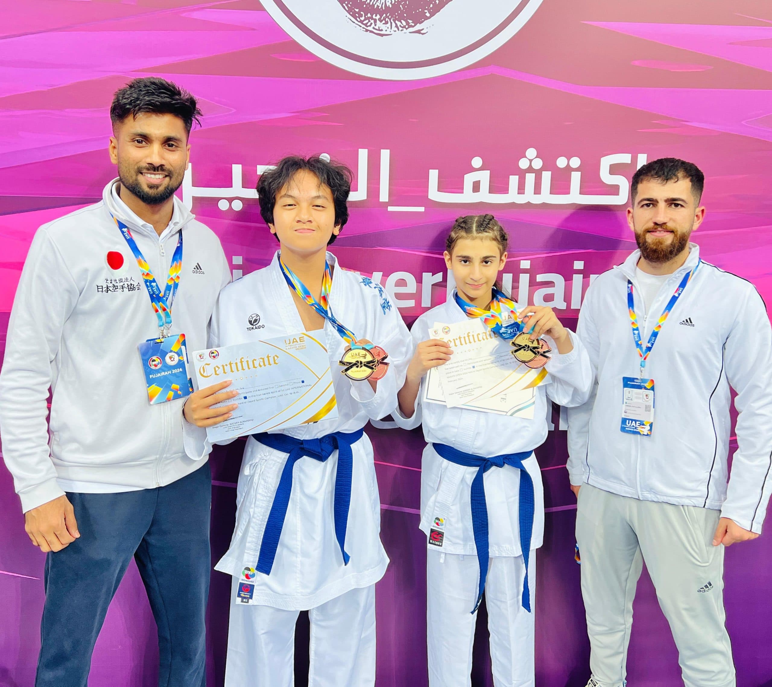 Two young medallists with certificates standing between their coaches at the Fujairah championship