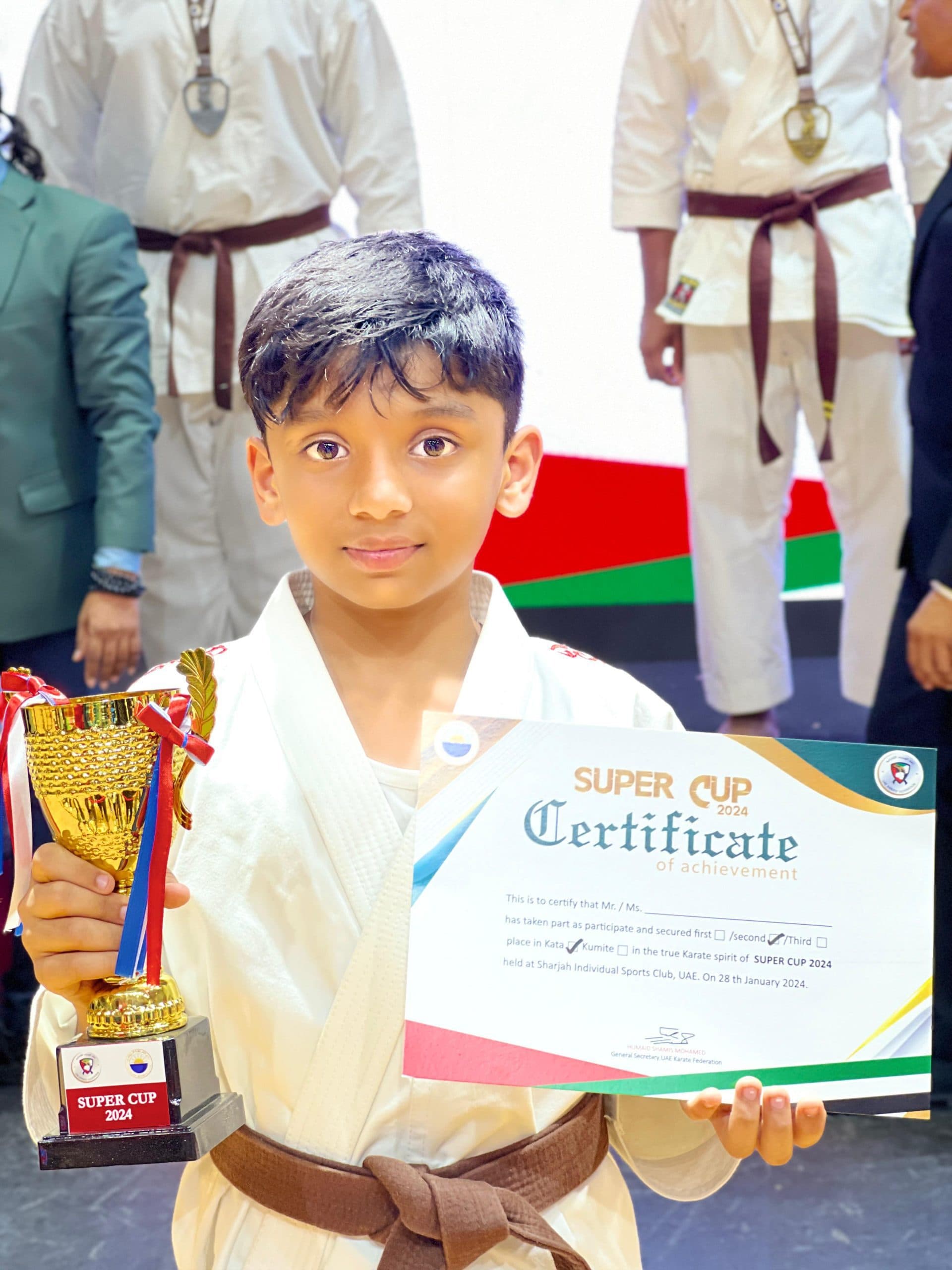 Young boy in white gi proudly holding a trophy and Super Cup certificate