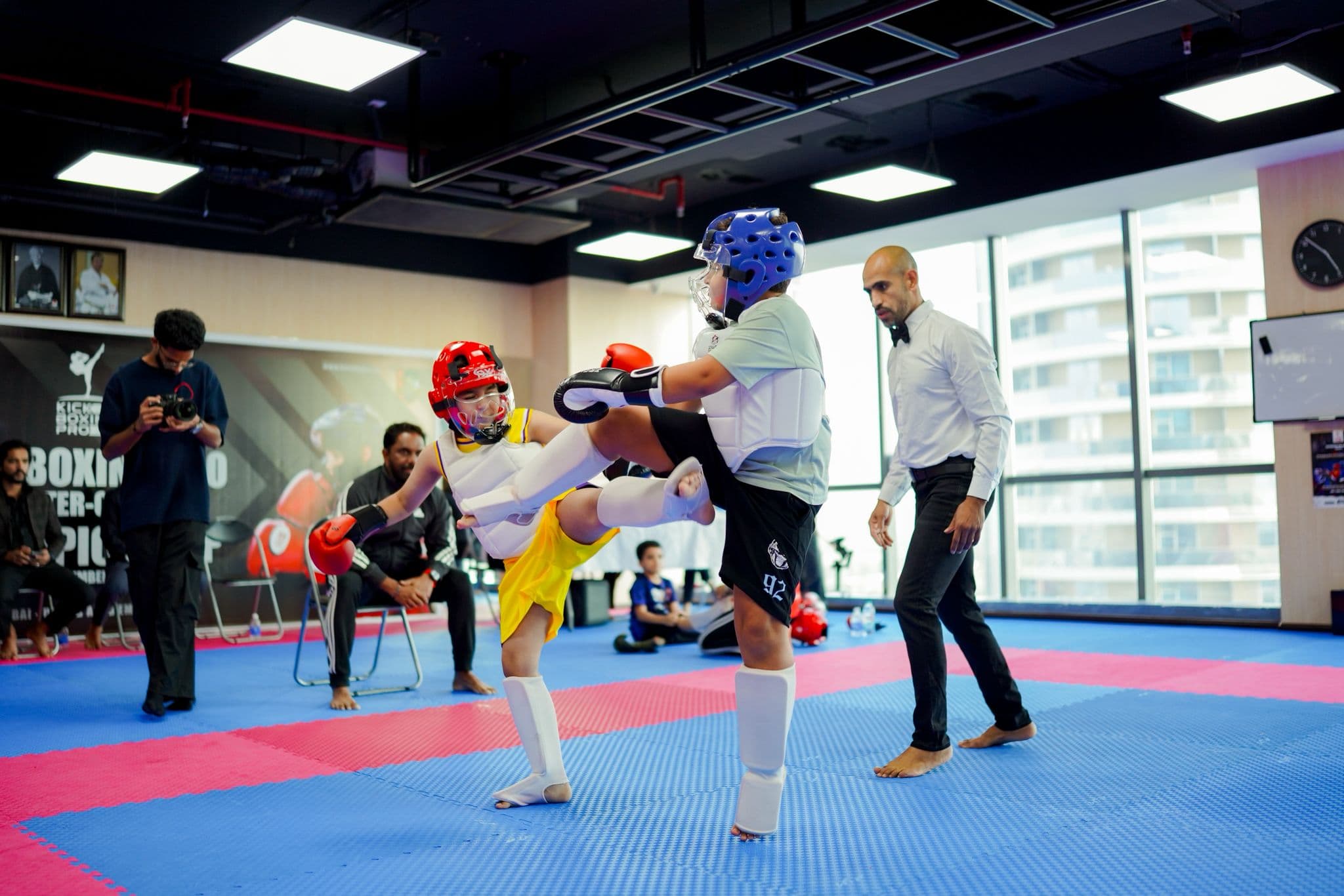 Kickboxer in a red head guard throwing a high kick during a bout by the dojo windows