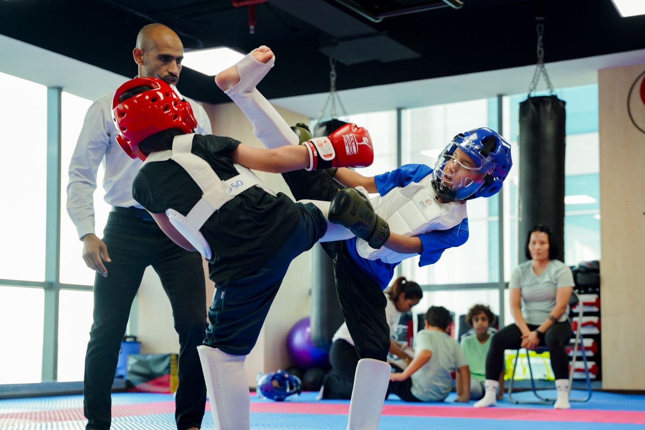 Young kickboxer in blue head guard landing a high kick on an opponent in red