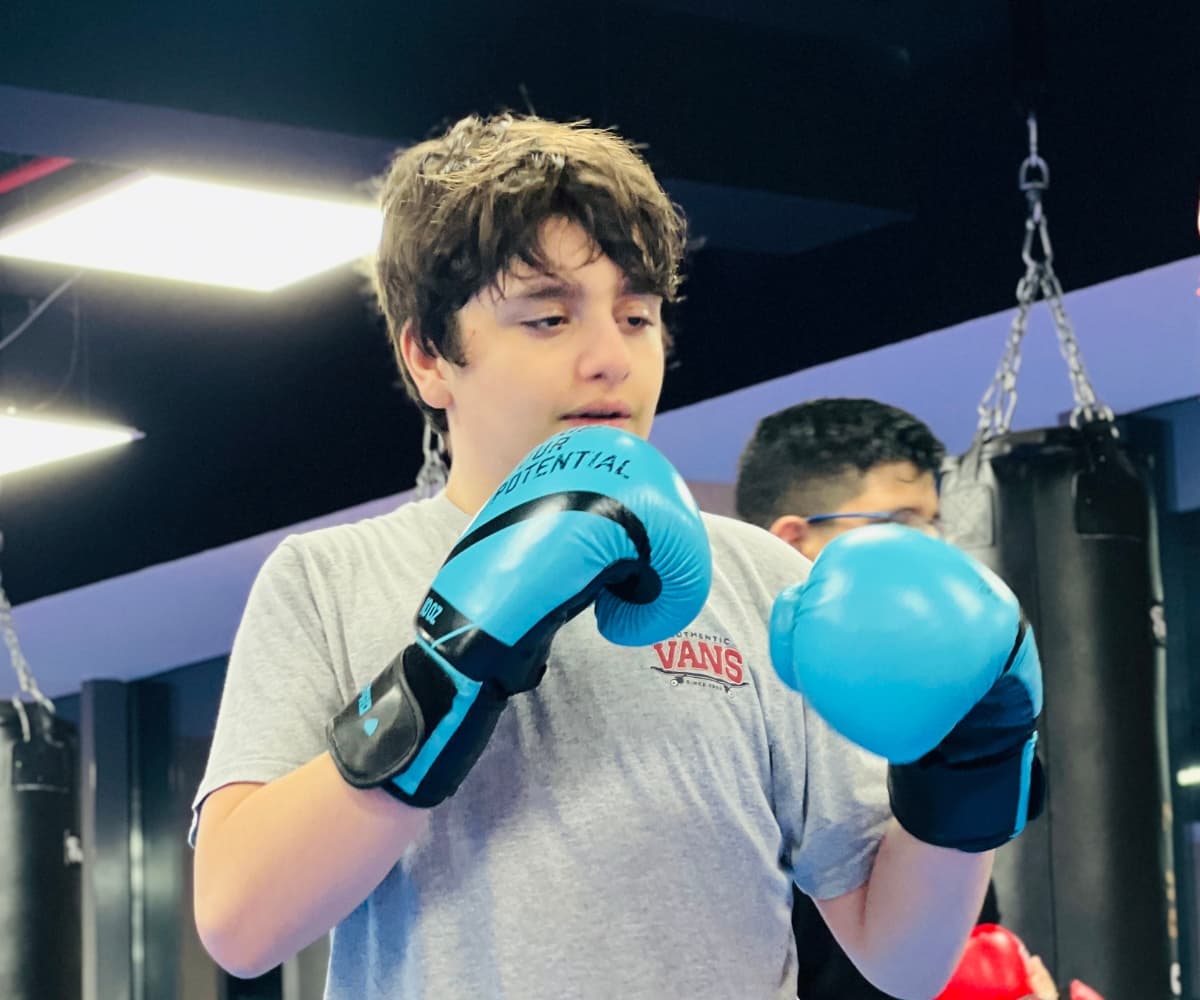 Teenager in blue boxing gloves holding a guard stance beside the heavy bags