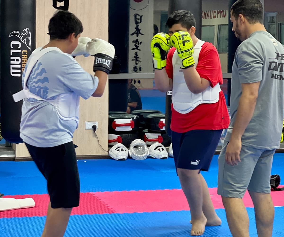 Two teenagers in body armour sparring with gloves and pads while a coach supervises