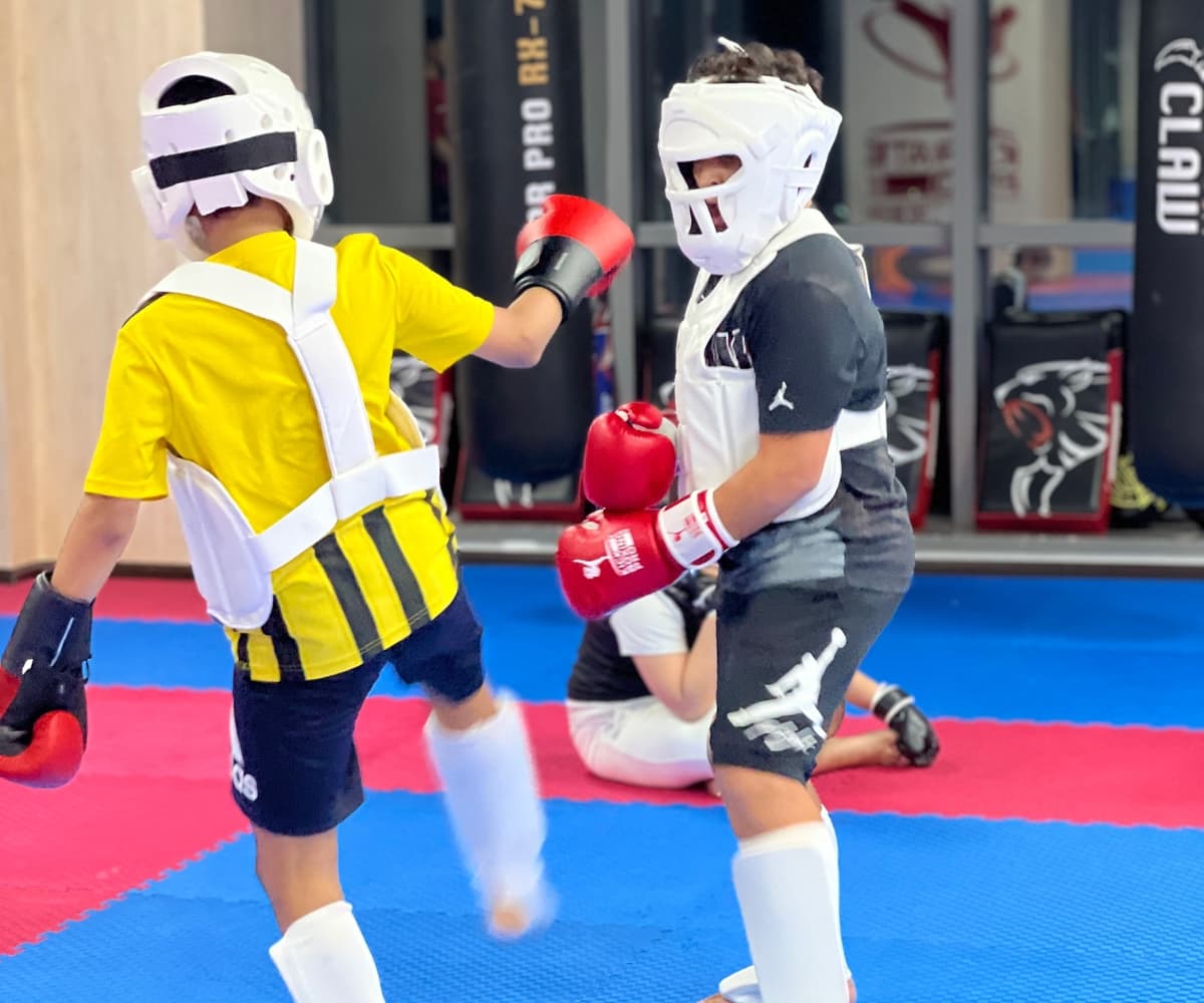 Two young kickboxers in head guards sparring on blue mats beside the heavy bags