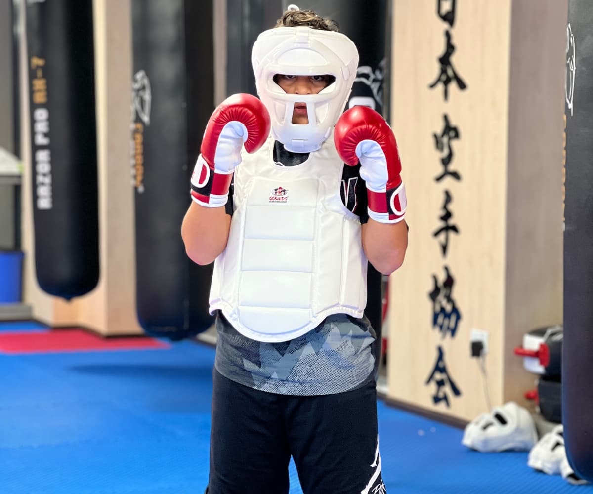 Young student in white head guard, body armour and red gloves posing under Japanese calligraphy