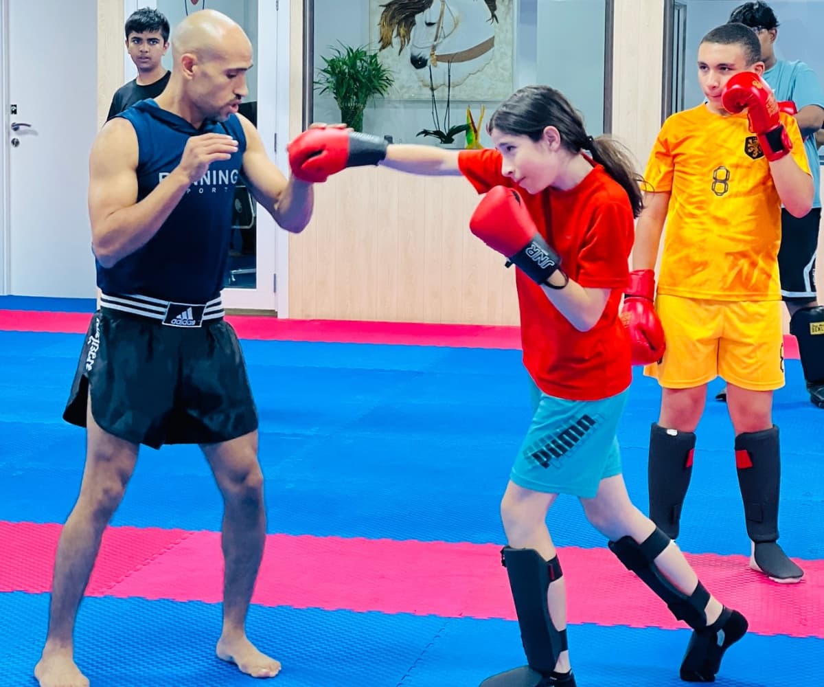 Coach holding a glove as a young girl in red practises her punch beside a classmate in yellow