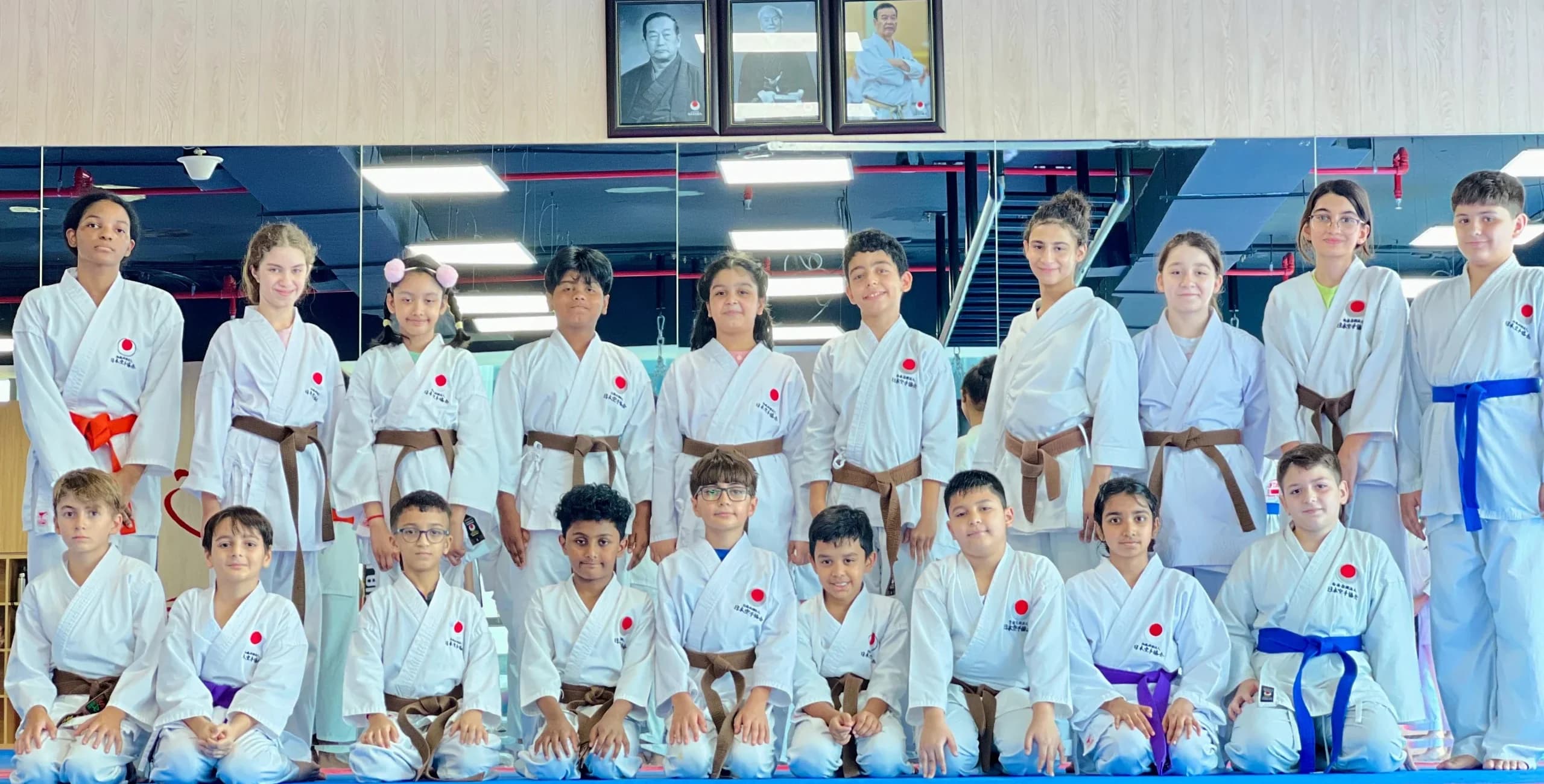 Kids karate class of mixed belt grades posing in rows beneath the dojo's framed portraits