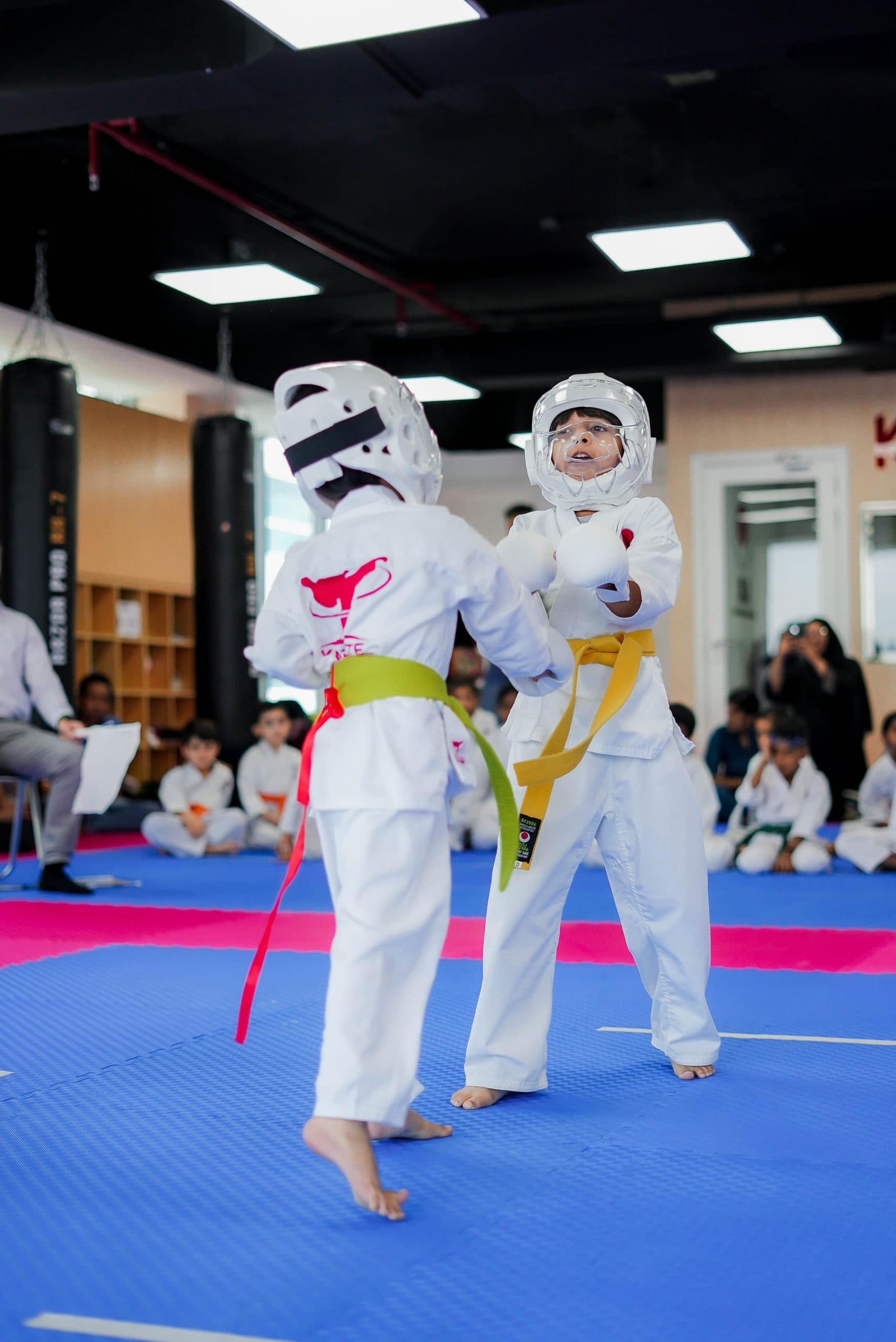 Two young students in head guards sparring while parents watch from the sidelines