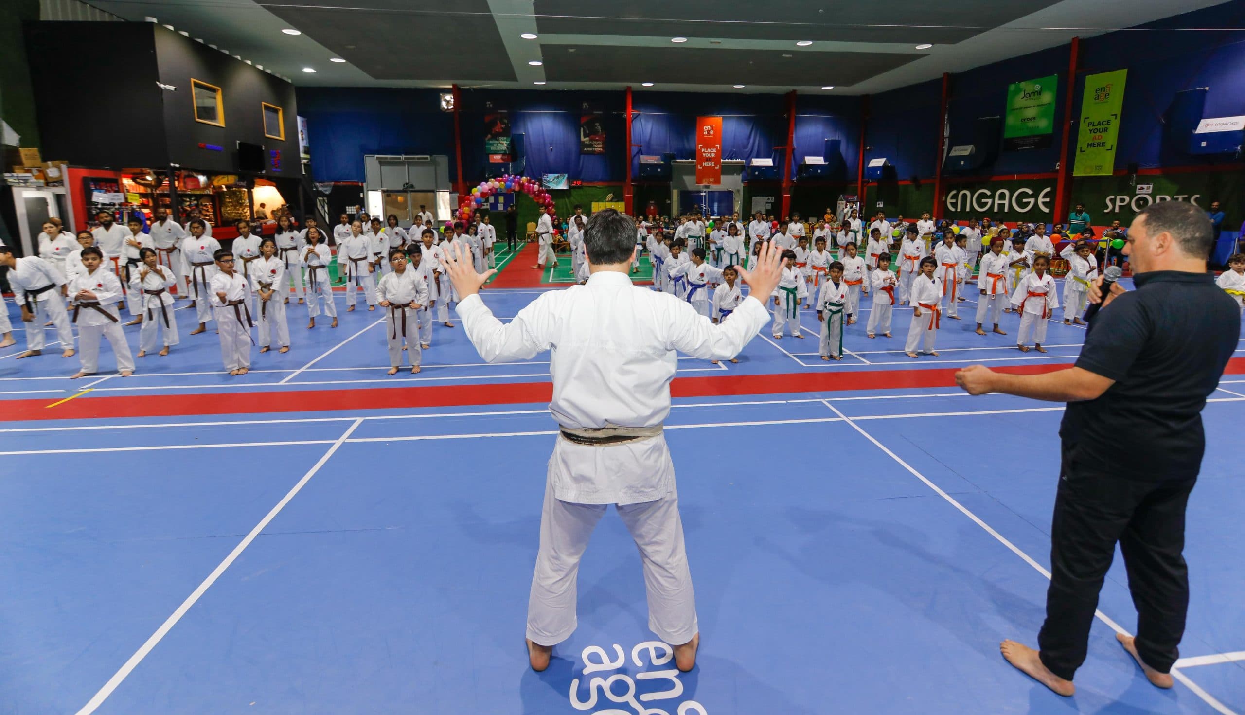Instructor in white gi leading a large group of children on a sports hall floor