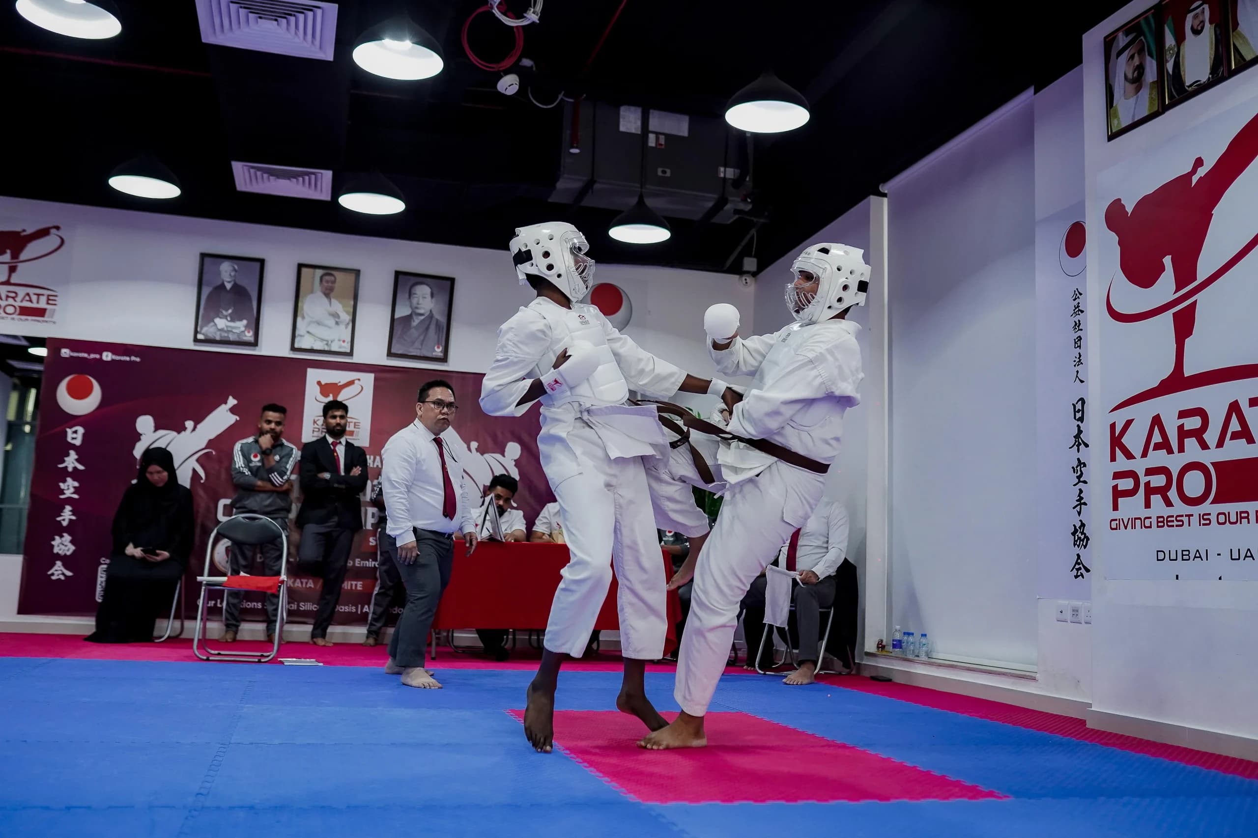 Two teenage karateka in protective headgear sparring in a kumite bout while judges watch